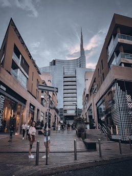 A modern urban scene with tall buildings flanking a pedestrian area. The central structure is a skyscraper with glass panels and a distinctive spire reaching into a cloudy sky. People are casually walking and interacting in the foreground, adding a lively touch to the architectural surroundings.