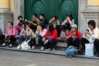 A group of people are seated on the steps in front of an ornate green door. They appear to be engaged with their phones and chatting, some with a disinterested expression. The individuals are dressed casually, with bags placed beside them.