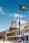 A colorful street lined with buildings features a prominent church tower with a clock. The sky is clear and blue, and the Bahamian flag flies on a pole. There are people walking and cars parked on the street.