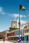 A colorful street lined with buildings features a prominent church tower with a clock. The sky is clear and blue, and the Bahamian flag flies on a pole. There are people walking and cars parked on the street.