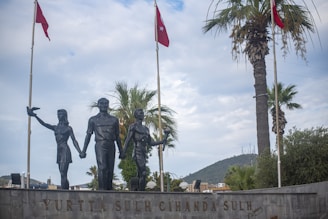 A statue depicts three figures holding hands, positioned in front of a backdrop of palm trees and flagpoles with red flags featuring a star and crescent. The scene is under a partly cloudy sky, with a distant view of a mountain and urban elements visible.