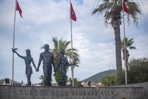 A statue depicts three figures holding hands, positioned in front of a backdrop of palm trees and flagpoles with red flags featuring a star and crescent. The scene is under a partly cloudy sky, with a distant view of a mountain and urban elements visible.