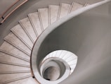 A stunning spiral staircase viewed from above, with a smooth curve and sleek design. The steps are made of stone, and the railing is wooden, creating an elegant and modern aesthetic.