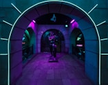 An arched hallway illuminated with vibrant neon lights features a statue at its center. The surrounding walls display a blend of glowing blue and purple hues, creating a futuristic and mysterious ambiance.