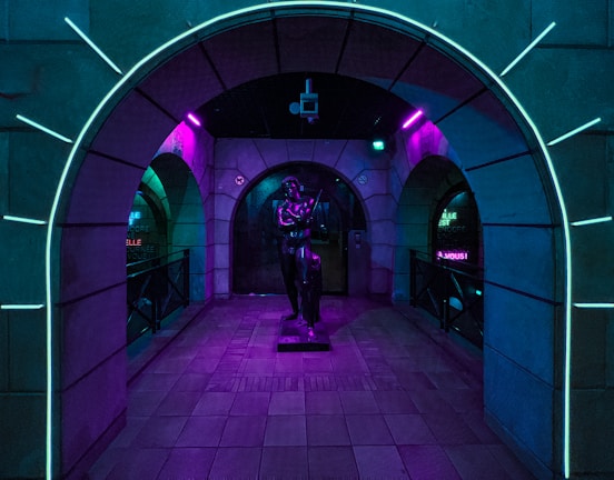 An arched hallway illuminated with vibrant neon lights features a statue at its center. The surrounding walls display a blend of glowing blue and purple hues, creating a futuristic and mysterious ambiance.