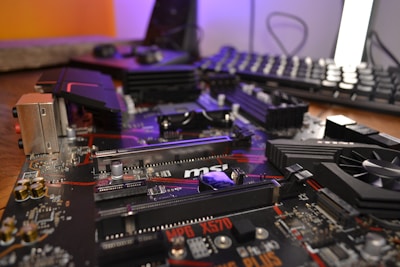 A close-up view of a complex computer motherboard with various electronic components including slots, heat sinks, and capacitors. The background features a blurred keyboard and other computer peripherals, hinting at a desktop setup.