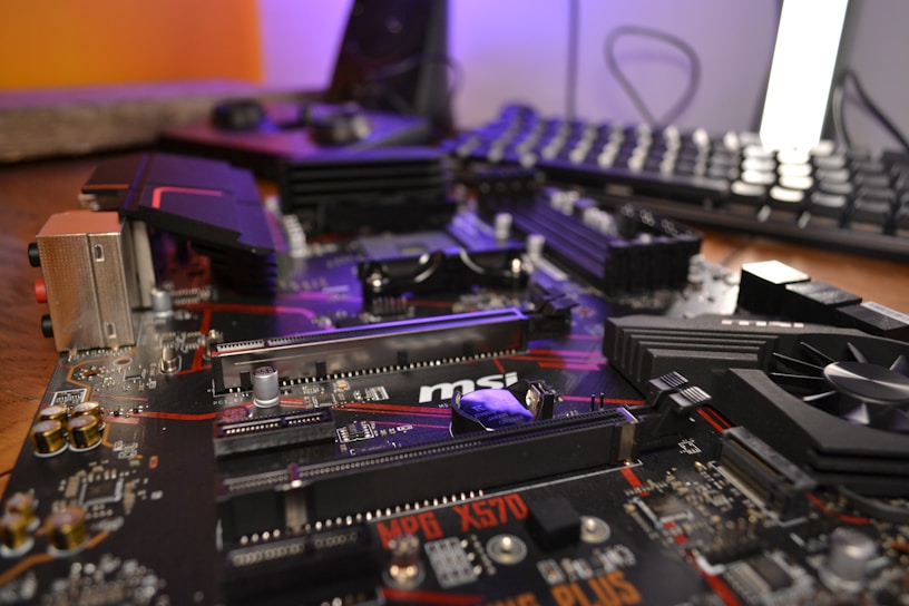 A close-up view of a complex computer motherboard with various electronic components including slots, heat sinks, and capacitors. The background features a blurred keyboard and other computer peripherals, hinting at a desktop setup.