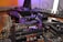 A close-up view of a complex computer motherboard with various electronic components including slots, heat sinks, and capacitors. The background features a blurred keyboard and other computer peripherals, hinting at a desktop setup.