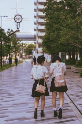 Two individuals wearing school uniforms walk along a tiled pathway bordered by trees in an urban area. Both carry backpacks and are engaged in conversation, with one wearing a pink backpack. Around them, there are buildings and a clock mounted on a streetlight.