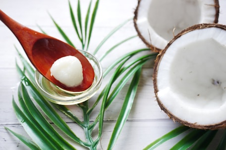 A wooden spoon holds a small portion of white coconut oil placed above a glass bowl, set on a lightly colored wooden surface. Two halved coconuts are visible, along with green palm fronds arranged decoratively.