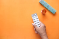 A hand holds a blister pack of tablets against an orange background. Nearby, a round orange pill bottle with tablets inside is open, and a weekly pill organizer is positioned with the compartments visible.