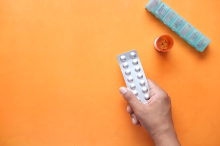 A hand holds a blister pack of tablets against an orange background. Nearby, a round orange pill bottle with tablets inside is open, and a weekly pill organizer is positioned with the compartments visible.