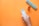 A hand holds a blister pack of tablets against an orange background. Nearby, a round orange pill bottle with tablets inside is open, and a weekly pill organizer is positioned with the compartments visible.