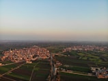 A sprawling aerial view of a rural area exhibiting a mix of densely packed buildings and expansive agricultural fields. A river or canal runs through the center of the image, diverging the town into two sections. Farmland fills the majority of the image, featuring various shades of green and brown, indicating different crop types or growth stages. The sky is mostly clear with a subtly warm glow from the setting or rising sun.