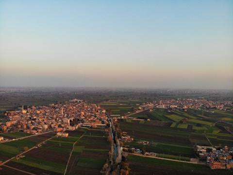 A sprawling aerial view of a rural area exhibiting a mix of densely packed buildings and expansive agricultural fields. A river or canal runs through the center of the image, diverging the town into two sections. Farmland fills the majority of the image, featuring various shades of green and brown, indicating different crop types or growth stages. The sky is mostly clear with a subtly warm glow from the setting or rising sun.