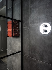 A modern bathroom interior featuring dark gray tiles on the walls. A sleek, black metal frame shelf holds a neatly stacked set of orange towels. A round chrome shower fixture is visible, contrasting with the textured wall surface.