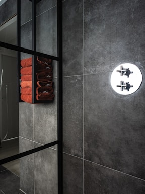 A modern bathroom interior featuring dark gray tiles on the walls. A sleek, black metal frame shelf holds a neatly stacked set of orange towels. A round chrome shower fixture is visible, contrasting with the textured wall surface.