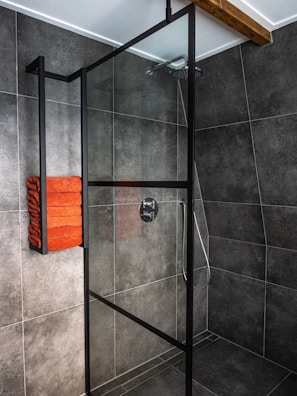 A modern bathroom features a spacious shower area with dark gray tiles covering the walls and floor. The shower is enclosed with a glass panel framed in black metal. Mounted on the wall is a set of neatly folded orange towels, adding a pop of color to the monochrome space. The showerhead and controls are sleek and contemporary, with a ceiling-mounted rain showerhead visible.