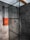 A modern bathroom features a spacious shower area with dark gray tiles covering the walls and floor. The shower is enclosed with a glass panel framed in black metal. Mounted on the wall is a set of neatly folded orange towels, adding a pop of color to the monochrome space. The showerhead and controls are sleek and contemporary, with a ceiling-mounted rain showerhead visible.
