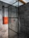 A modern bathroom features a spacious shower area with dark gray tiles covering the walls and floor. The shower is enclosed with a glass panel framed in black metal. Mounted on the wall is a set of neatly folded orange towels, adding a pop of color to the monochrome space. The showerhead and controls are sleek and contemporary, with a ceiling-mounted rain showerhead visible.