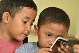 Two young children are closely examining a smartphone, with expressions of concentration and curiosity. They appear to be deeply engaged with what is on the screen.