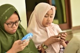 Two young girls are intently focused on their smartphones. They are sitting side by side, with one wearing a pink hijab and the other wearing a green hijab and glasses. The setting appears to be indoors, possibly a hall or waiting area.