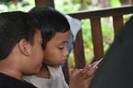 Two children are closely seated and focusing intently on a smartphone, with one child resting on the other's shoulder. They are in a wooden structure, possibly a porch, with greenery visible outside.