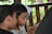 Two children are closely seated and focusing intently on a smartphone, with one child resting on the other's shoulder. They are in a wooden structure, possibly a porch, with greenery visible outside.