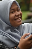 A person is smiling and looking off to the side while holding a phone. They are wearing a light blue hijab and appear to be outdoors with a blurred green background.
