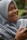 A person is smiling and looking off to the side while holding a phone. They are wearing a light blue hijab and appear to be outdoors with a blurred green background.