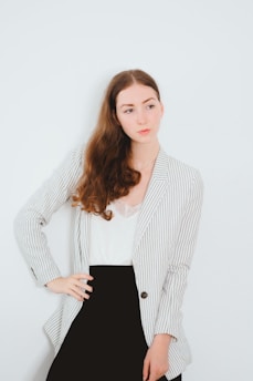 A woman with long brown hair is standing against a plain white background, wearing a white blouse, a striped blazer, and a black skirt. Her expression is neutral, and she has one hand on her hip.