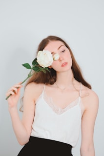 A young woman with long hair holds a white rose in front of her face. She is wearing a white lace camisole and a black skirt, with a minimalistic necklace. Her expression is serene, and she is positioned against a plain light blue background.
