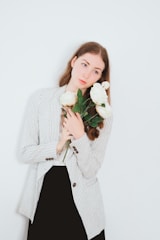 A person with long brown hair holds a bouquet of large white roses, wearing a striped blazer over a black dress. The background is plain light blue.