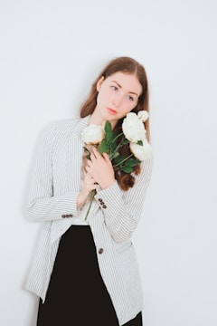 A person with long brown hair holds a bouquet of large white roses, wearing a striped blazer over a black dress. The background is plain light blue.