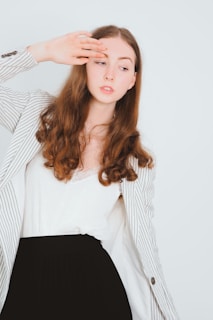A young woman with long, curly hair gazes sideways, wearing a white top and a striped blazer. Her hand is raised near her forehead, as if shielding her eyes from light.