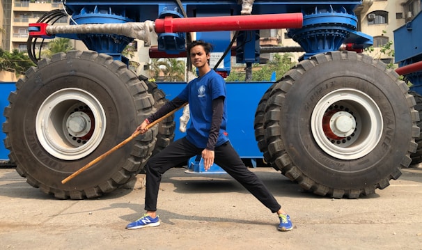 A person wearing a blue shirt and black pants stands in a martial arts pose with a wooden staff. They are positioned in front of a large blue vehicle with two massive off-road tires. The background shows a few buildings and palm trees.