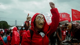 A person wearing a Salvador Dalí mask and a red hooded outfit raises a fist in a crowd. Many individuals are also dressed in red clothing and some carry red flags with inscriptions. The scene suggests a demonstration or public gathering, with various people wearing masks in the background.