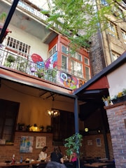An outdoor seating area of a café is visible, with people dining at tables. The setting is surrounded by brick walls, with plants and colorful artwork decorating the space. Overhead, a balcony with vibrant street art and potted plants adds character to the scene. The mood is laid-back and inviting, enhanced by a chandelier lighting fixture.
