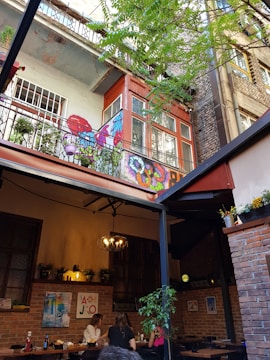 An outdoor seating area of a café is visible, with people dining at tables. The setting is surrounded by brick walls, with plants and colorful artwork decorating the space. Overhead, a balcony with vibrant street art and potted plants adds character to the scene. The mood is laid-back and inviting, enhanced by a chandelier lighting fixture.