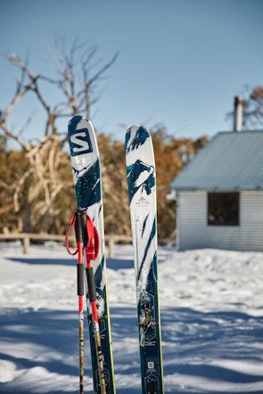 Close-up of ski boots and poles resting on snow beside a mountain lodge.