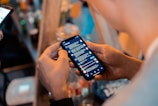 A person holding a smartphone with a messaging app open, showing a list of conversations. Another device is visible in the background, and there are blurred bottles and objects on a table in a dimly lit setting, suggesting a casual or social environment.