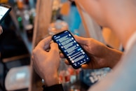 A person holding a smartphone with a messaging app open, showing a list of conversations. Another device is visible in the background, and there are blurred bottles and objects on a table in a dimly lit setting, suggesting a casual or social environment.