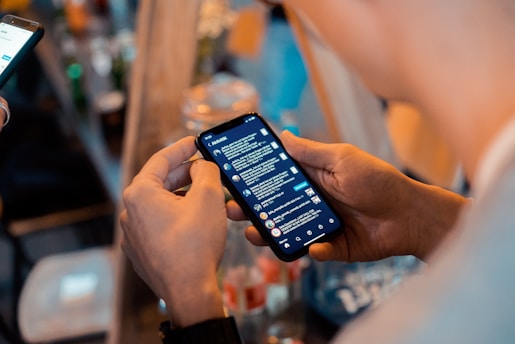 A person holding a smartphone with a messaging app open, showing a list of conversations. Another device is visible in the background, and there are blurred bottles and objects on a table in a dimly lit setting, suggesting a casual or social environment.