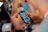 A person holding a smartphone with a messaging app open, showing a list of conversations. Another device is visible in the background, and there are blurred bottles and objects on a table in a dimly lit setting, suggesting a casual or social environment.