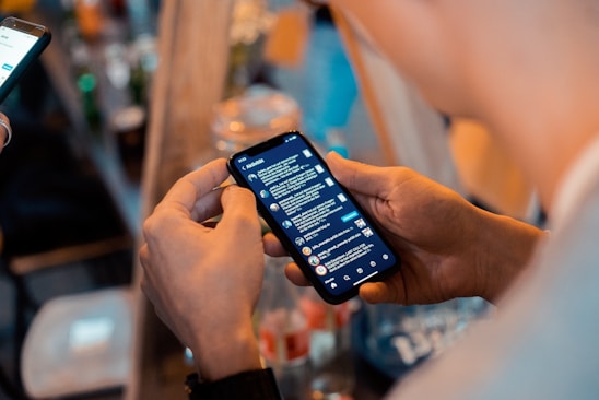 A person holding a smartphone with a messaging app open, showing a list of conversations. Another device is visible in the background, and there are blurred bottles and objects on a table in a dimly lit setting, suggesting a casual or social environment.