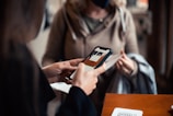 Two people are engaged in a transaction, with the focus on a hand holding a smartphone displaying an application. The background shows another person wearing a mask and holding a piece of clothing, suggesting an indoor setting.