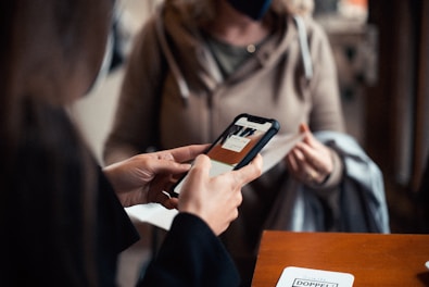 Two people are engaged in a transaction, with the focus on a hand holding a smartphone displaying an application. The background shows another person wearing a mask and holding a piece of clothing, suggesting an indoor setting.