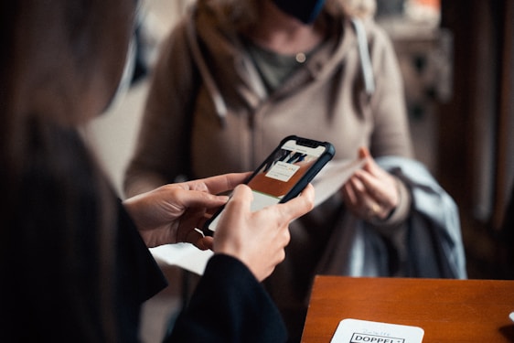 Two people are engaged in a transaction, with the focus on a hand holding a smartphone displaying an application. The background shows another person wearing a mask and holding a piece of clothing, suggesting an indoor setting.