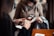 Two people are engaged in a transaction, with the focus on a hand holding a smartphone displaying an application. The background shows another person wearing a mask and holding a piece of clothing, suggesting an indoor setting.
