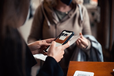 Two people are engaged in a transaction, with the focus on a hand holding a smartphone displaying an application. The background shows another person wearing a mask and holding a piece of clothing, suggesting an indoor setting.